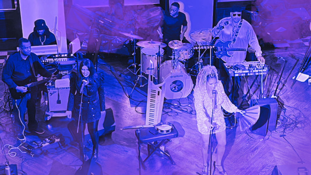 Full live band performing on stage with lead singer, Joy Stom, backup vocals, drums, keyboard, and guitar players